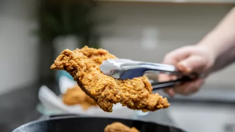 fried chicken being held in the air with metal tongs over an iron skillet