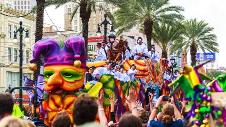 	crowds celebrating Mardi Gras as a float makes its way along the street. men in white and masks throwing beads into the crowds.