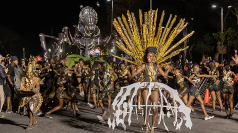 	a parade of people in black and yellow costumes lead by a woman in an elaborate costume followed by a giant ant sculpture