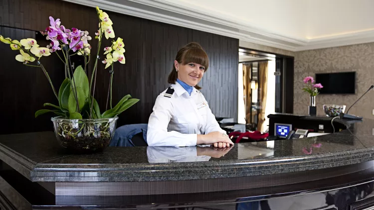 Internal shot of woman in uniform politely smiling behind a glossy, black reception desk inside the William Shakespeare cruise ship