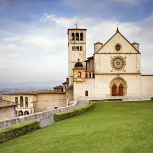 The beautiful Basilica of St. Francis of Assisi located in the town of Assisi, Italy. Photo contains a Tuscan hillside view in the background and a bright blue sky with pretty cloud formations.