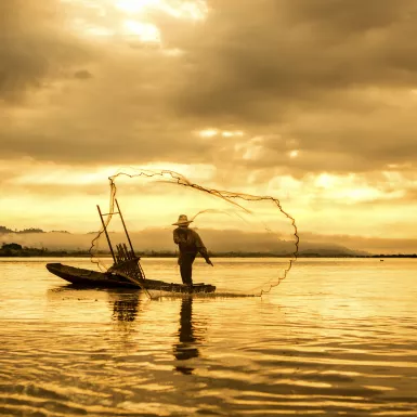 A fisherman in a boat throwing a net on the lake during sunset