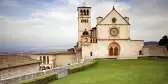 The beautiful Basilica of St. Francis of Assisi located in the town of Assisi, Italy. Photo contains a Tuscan hillside view in the background and a bright blue sky with pretty cloud formations.