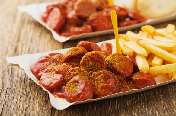 Traditional German currywurst, served with chips on disposable paper tray. Wooden table as background.