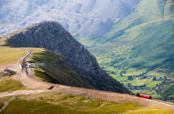 A train descends from the summit of Snowdon Mountain on a narrow railway with beautiful scenery in the background.