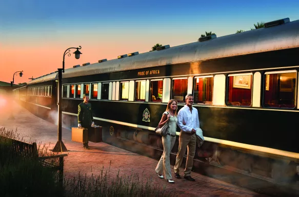 Mature couple walking on platform beside the Pride of Africa train and a man carrying luggage