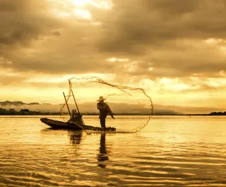 A fisherman in a boat throwing a net on the lake during sunset