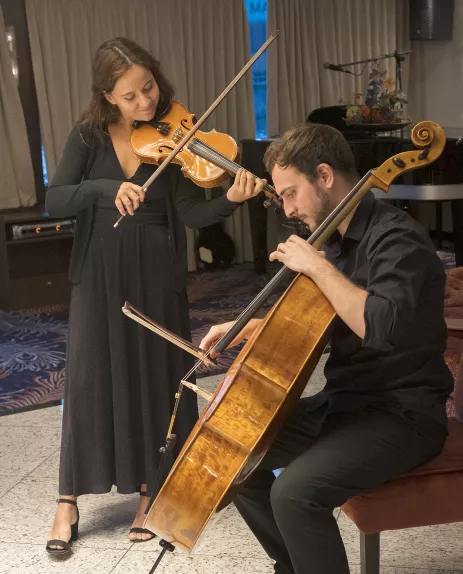 Two formally clothed musician performing, one standing playing the violin and one seated playing the cello  